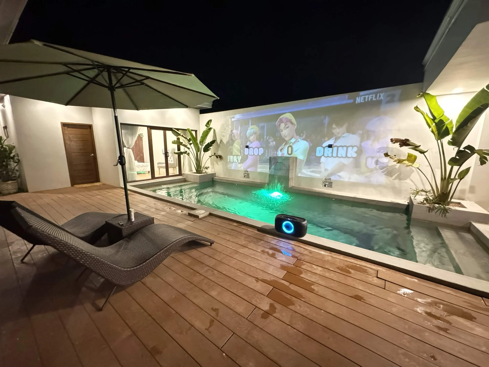 Pool deck transformed into outdoor cinema at night with projector, screen on the wall and underwater pool lighting